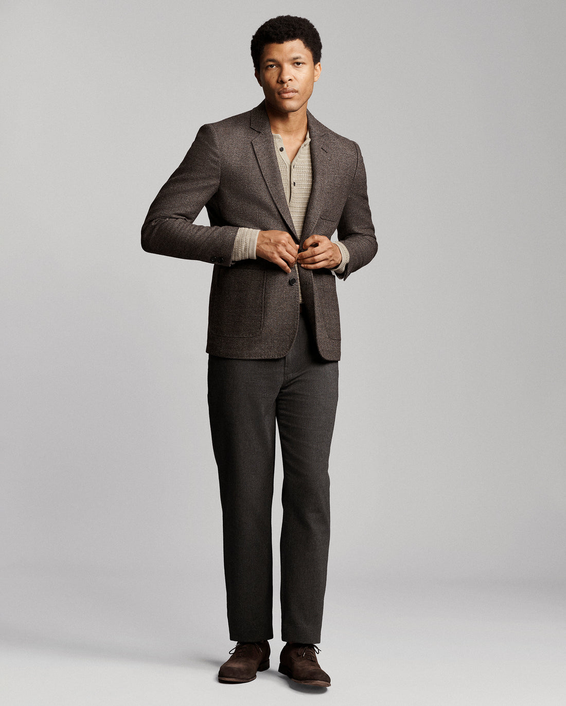 A man modeling a dark gray two-piece suit with a textured blazer and matching trousers, styled over a beige knit shirt and brown dress shoes, against a light gray studio background.