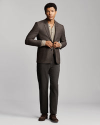 A man modeling a dark gray two-piece suit with a textured blazer and matching trousers, styled over a beige knit shirt and brown dress shoes, against a light gray studio background.