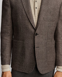 A close-up of a men's brown tweed blazer with notch lapels, a two-button closure, and front patch pockets, styled over a cream knit sweater and gray trousers.