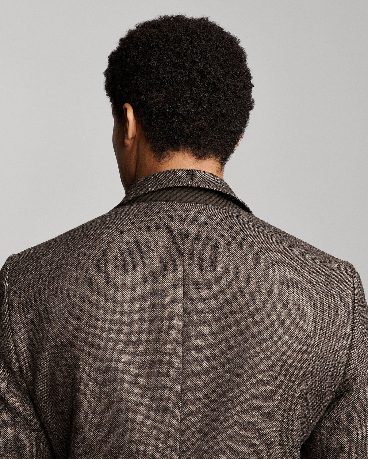 Rear view of a man wearing a textured brown herringbone tweed blazer, showing the back collar and shoulder details against a solid gray background.
