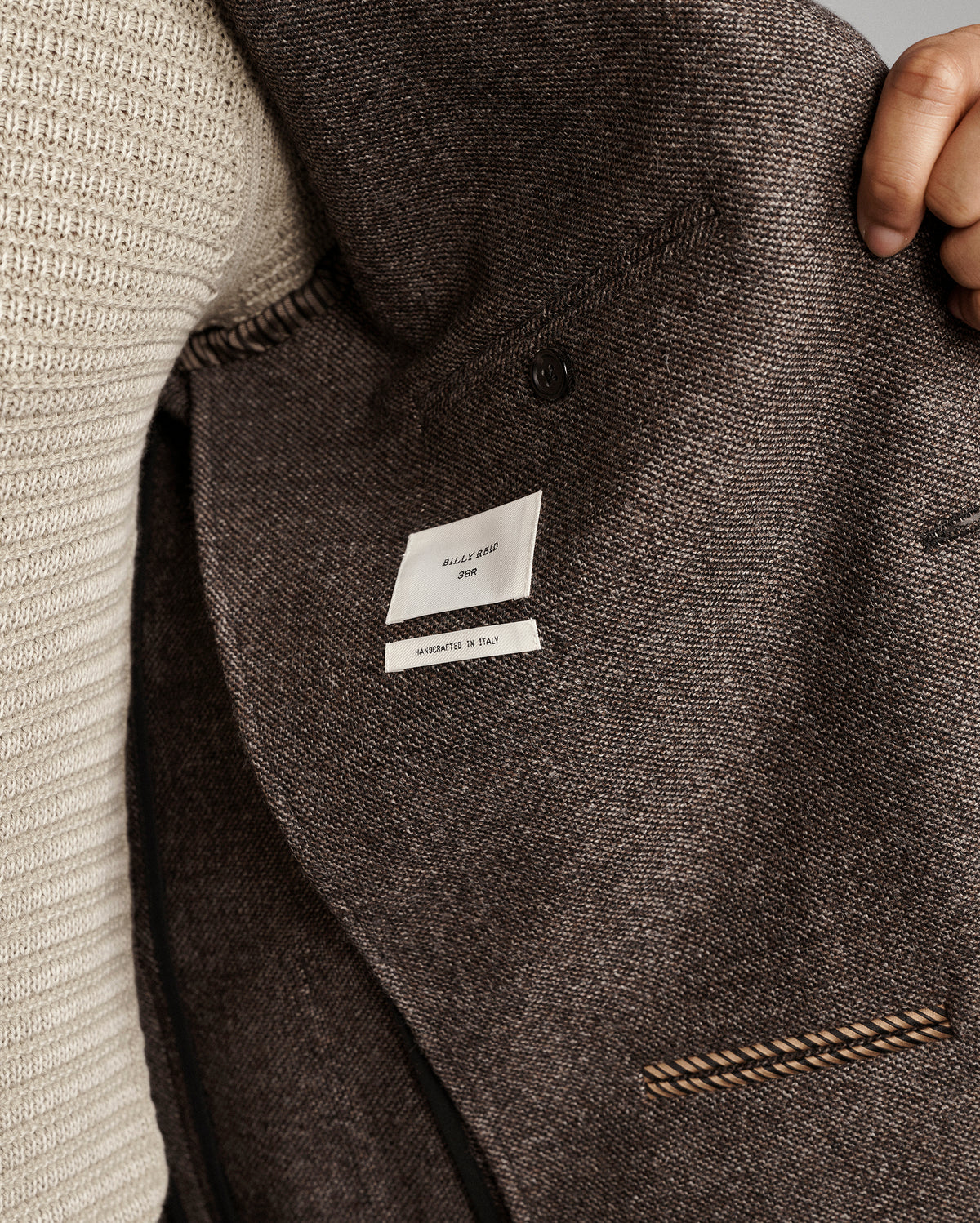 Close-up of a dark brown tweed blazer jacket with a white brand label reading 'BILLY REID' and 'Made in Italy', worn over a cream ribbed knit sweater, with a hand holding the collar.
