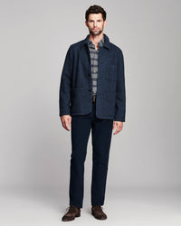 A man modeling a dark navy blue textured jacket over a gray and white plaid shirt, paired with navy trousers and brown leather shoes, against a light gray studio background.