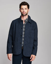 A man wearing a dark navy blue textured jacket over a gray patterned shirt, standing against a solid gray studio background.
