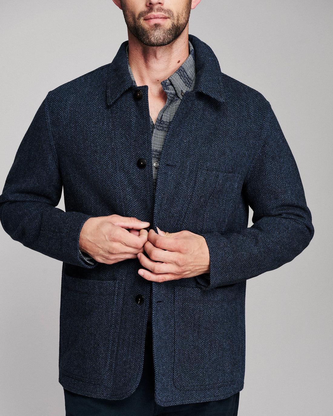 A man wearing a dark navy blue textured wool-blend jacket over a gray plaid shirt, shown from the chin down against a solid gray studio background.