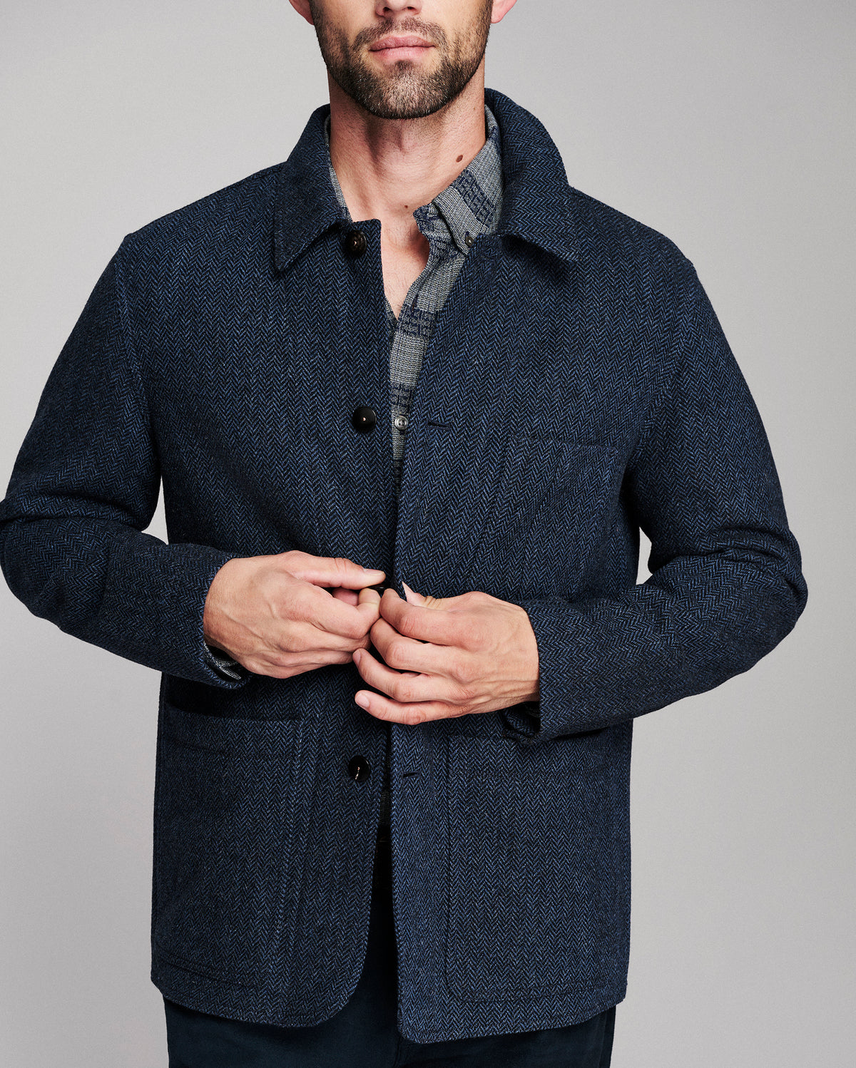 A man wearing a dark navy blue textured wool-blend jacket over a gray plaid shirt, shown from the chin down against a solid gray studio background.