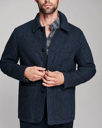 A man wearing a dark navy blue textured wool-blend jacket over a gray plaid shirt, shown from the chin down against a solid gray studio background.