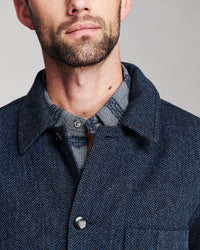 Close-up of a man wearing a dark navy textured overshirt over a blue and gray plaid button-down shirt, against a neutral gray background.