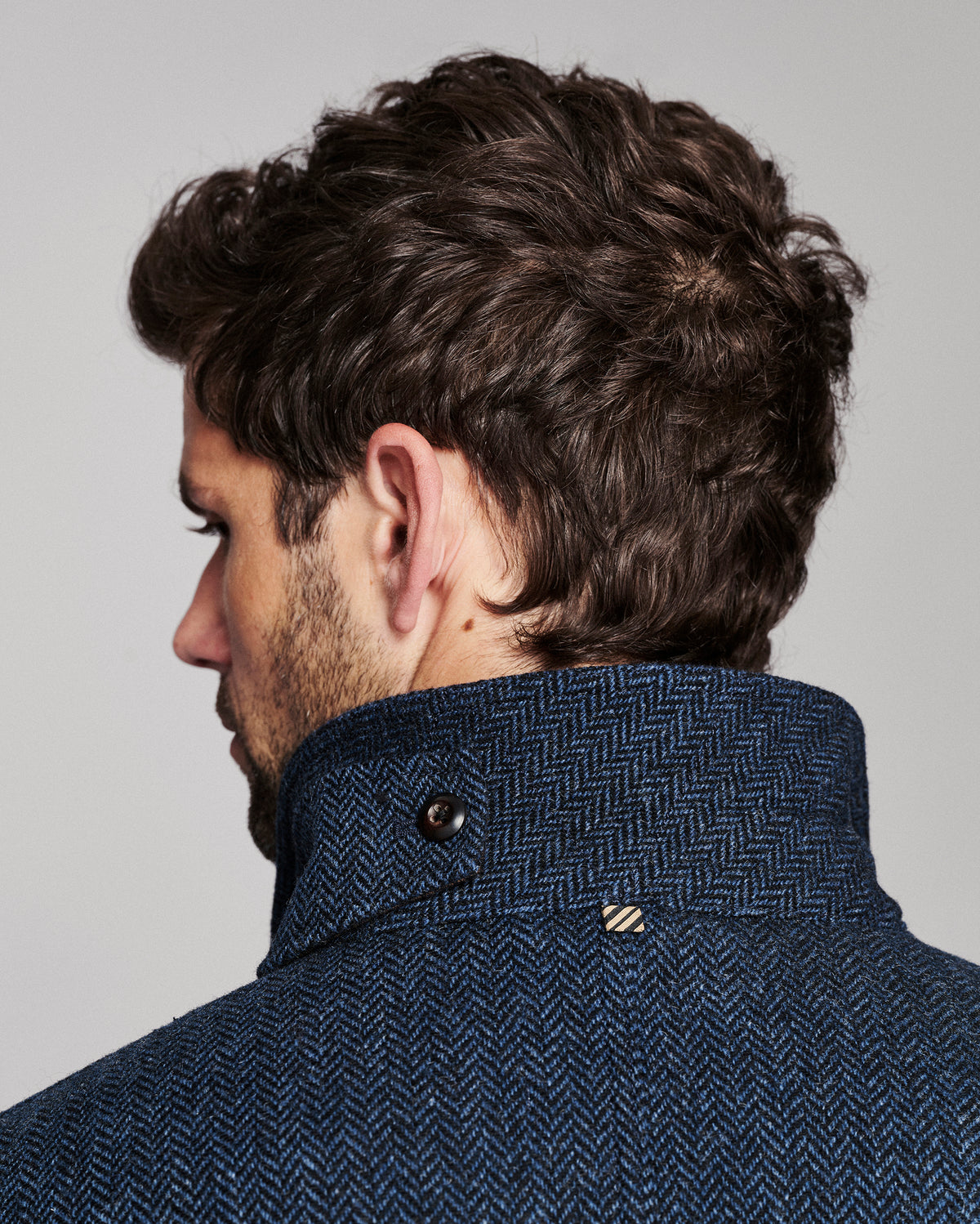 Close-up side profile of a man wearing a textured navy blue herringbone jacket with a high buttoned collar, against a light gray studio background.