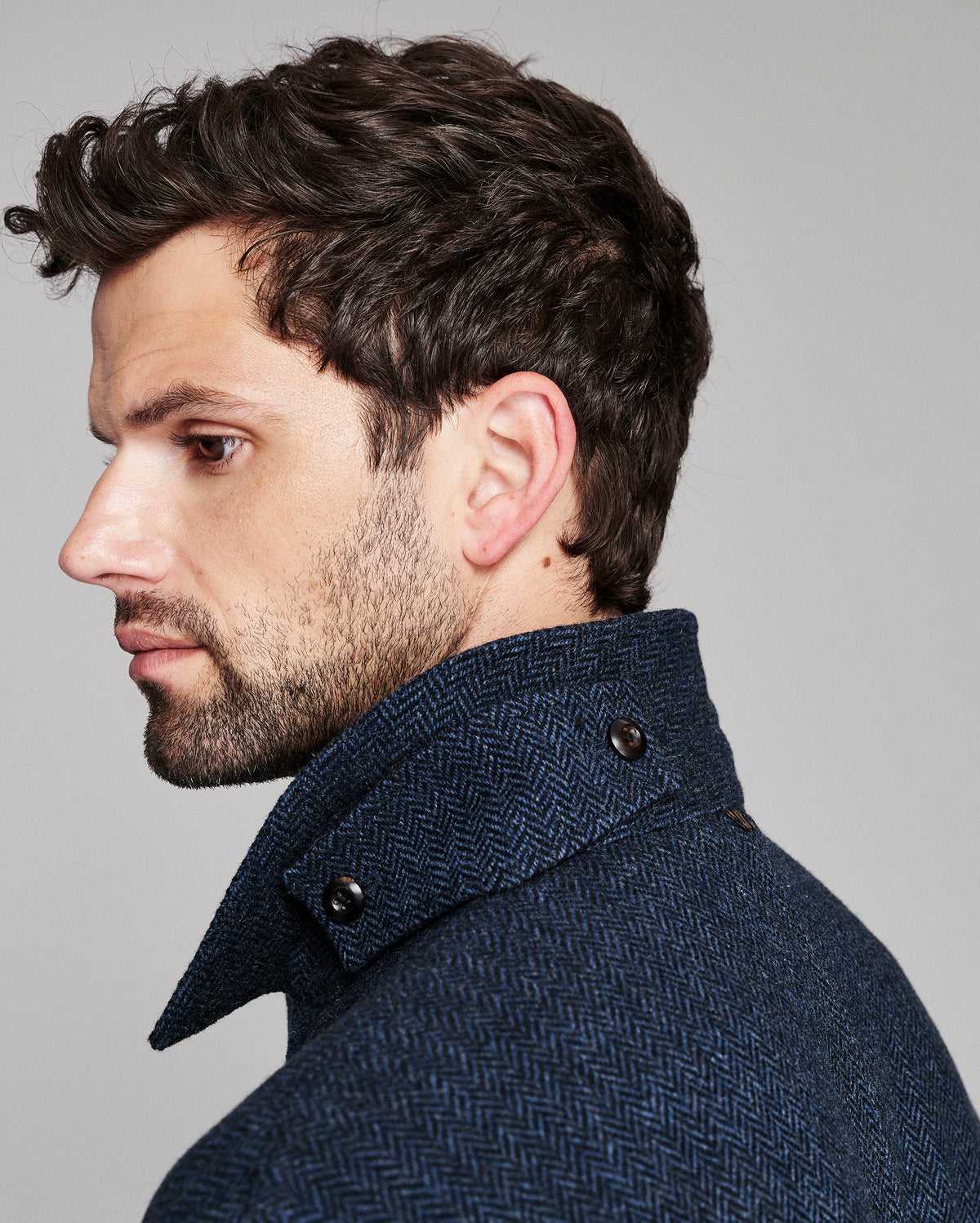 A close-up side profile of a man wearing a textured navy blue herringbone coat with a high structured collar, set against a solid light gray background.