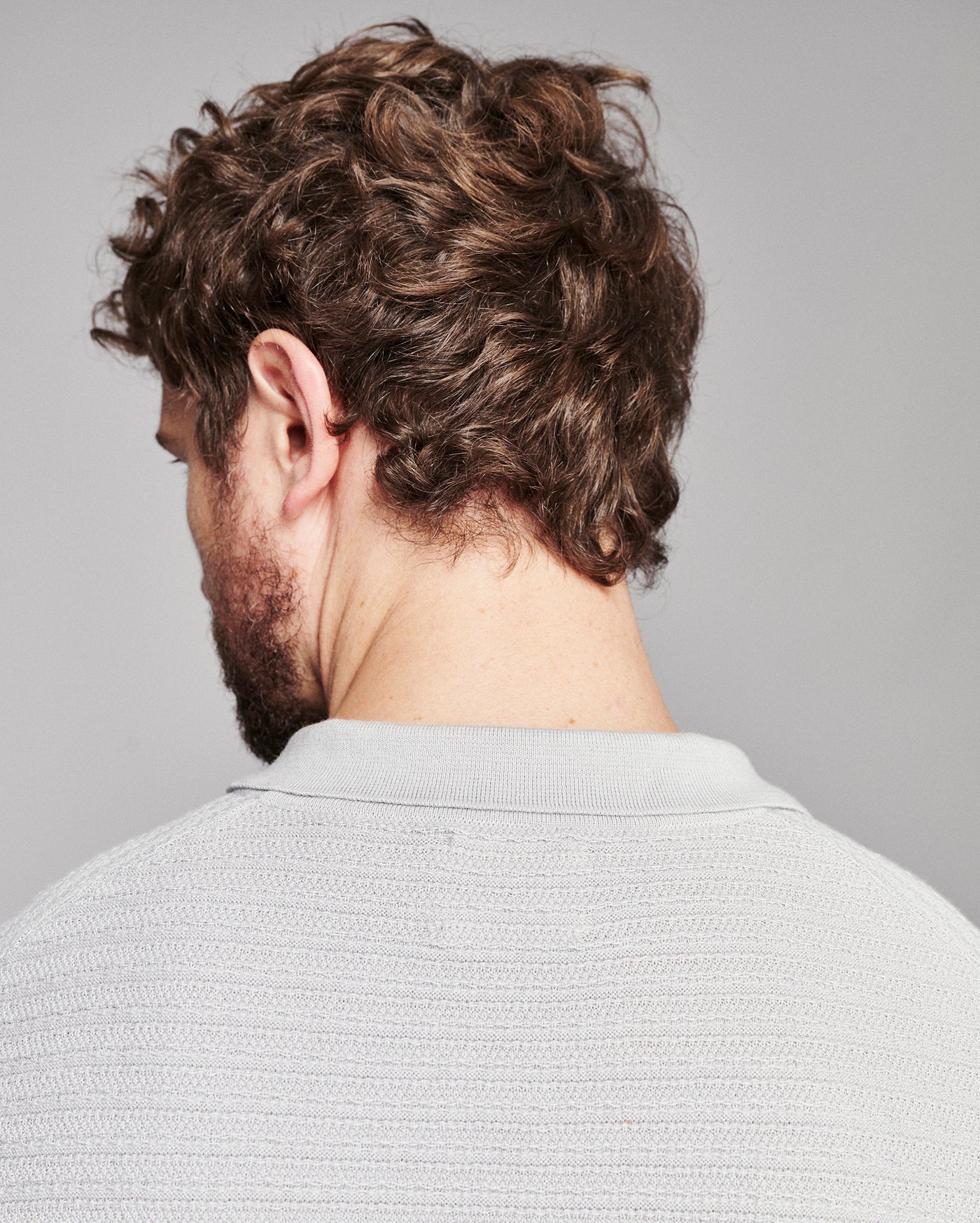 Rear view of a man with curly brown hair and a beard, wearing a light gray textured knit polo shirt against a solid gray background.