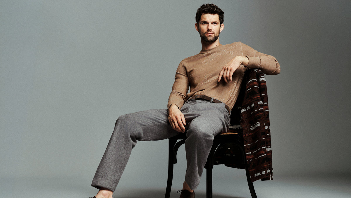 A man seated on a dark chair against a plain gray studio background, wearing a fitted tan long-sleeve top, light gray trousers, and dark shoes, with a patterned brown-and-red draped textile over his right shoulder.