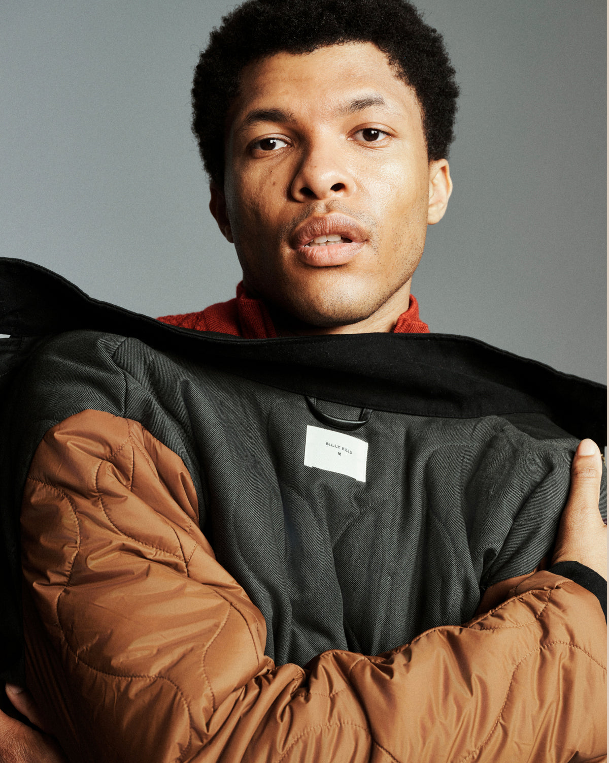 A close-up portrait of a man wearing a black and brown puffer jacket, holding it open to show the interior lining. The jacket features a black outer shell and quilted brown sleeves and collar.
