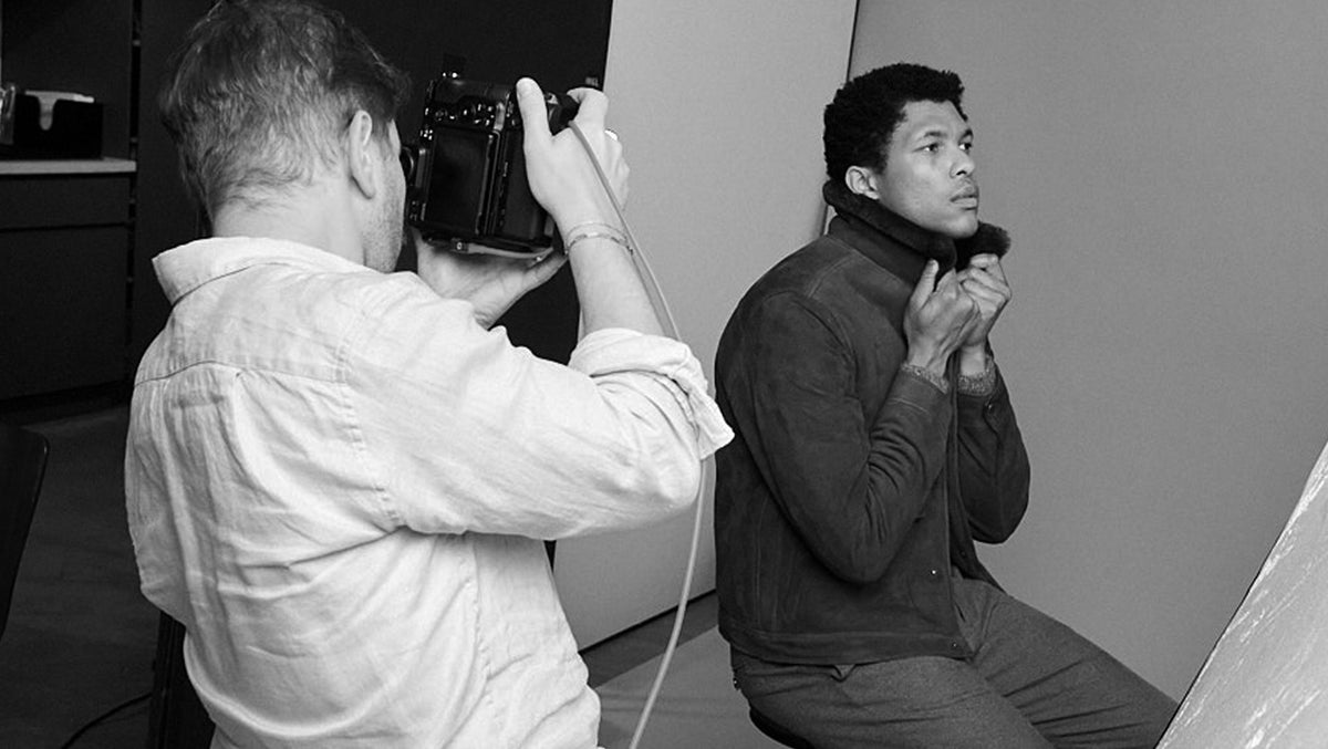 A black and white photograph showing a photographer holding a camera to shoot a model wearing a dark turtleneck sweater in a studio setting.