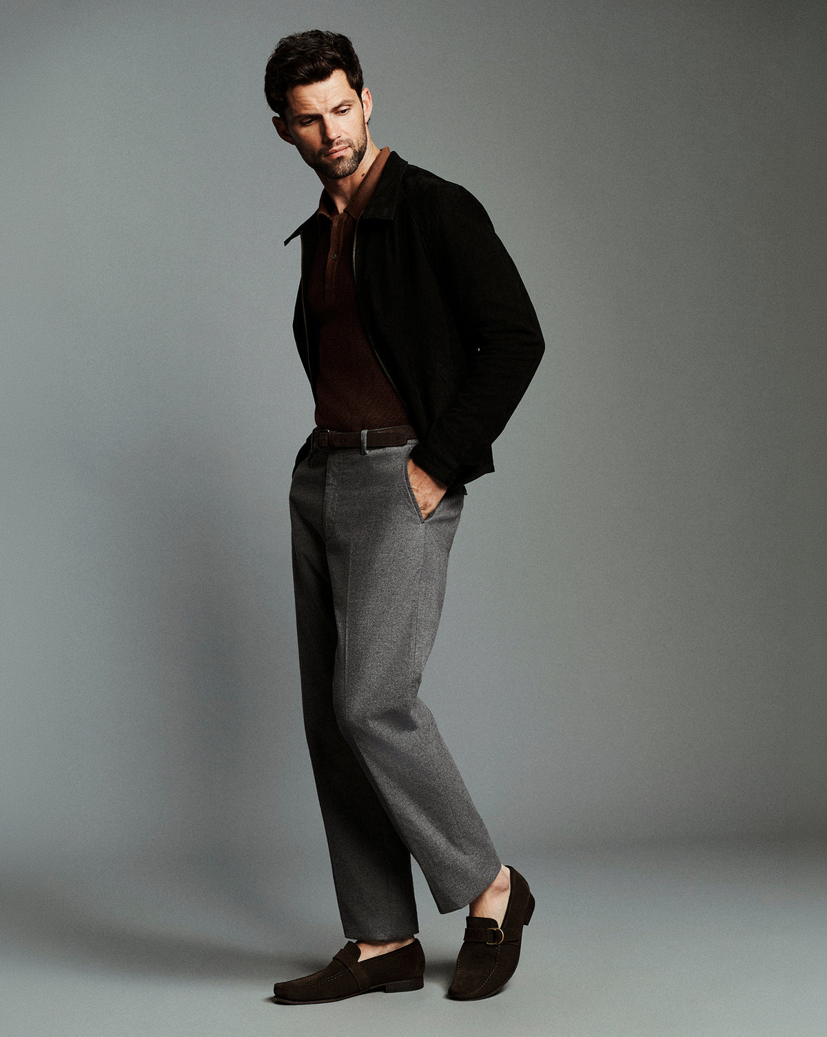 A man modeling a dark brown polo shirt under a black jacket, paired with gray trousers and dark brown loafers, against a solid gray studio background.