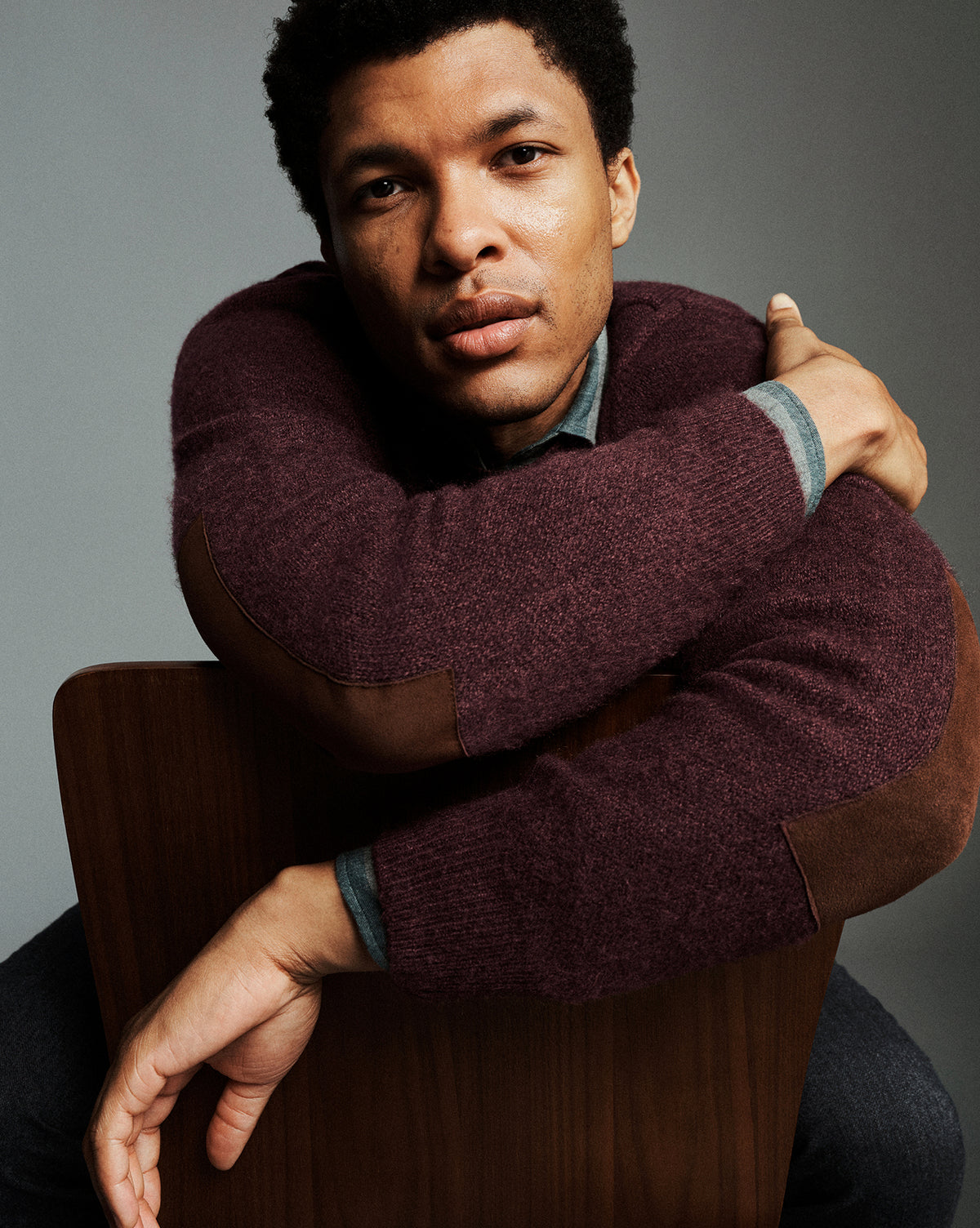 A man wearing a burgundy knit sweater with brown elbow patches and a blue collared shirt underneath, leaning on a wooden chair against a gray studio background.