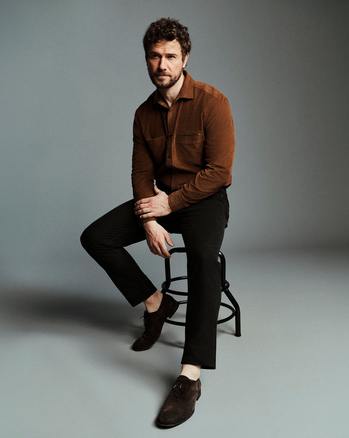 A man with curly brown hair and a beard sits on a black metal stool, wearing a brown corduroy button-up shirt, black trousers, and dark brown suede loafers against a solid gray studio background.