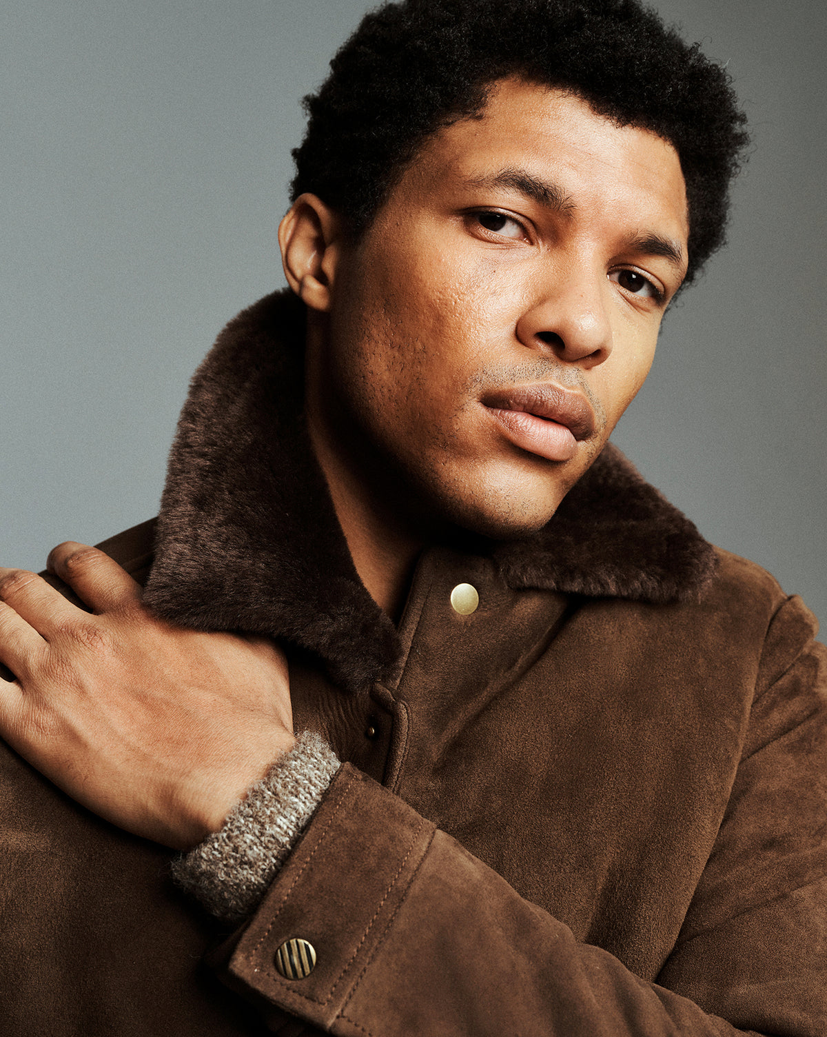 A close-up portrait of a man wearing a brown suede jacket with a dark brown faux-fur collar, set against a solid gray studio background.