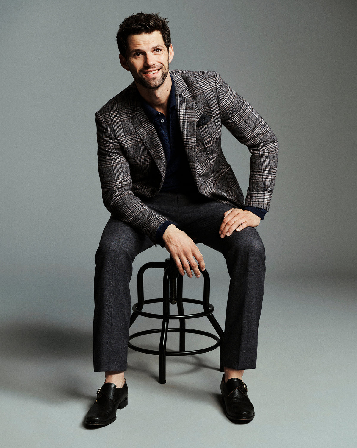 A man wearing a gray plaid blazer, dark trousers, and black loafers sits on a black metal stool against a solid gray studio background.