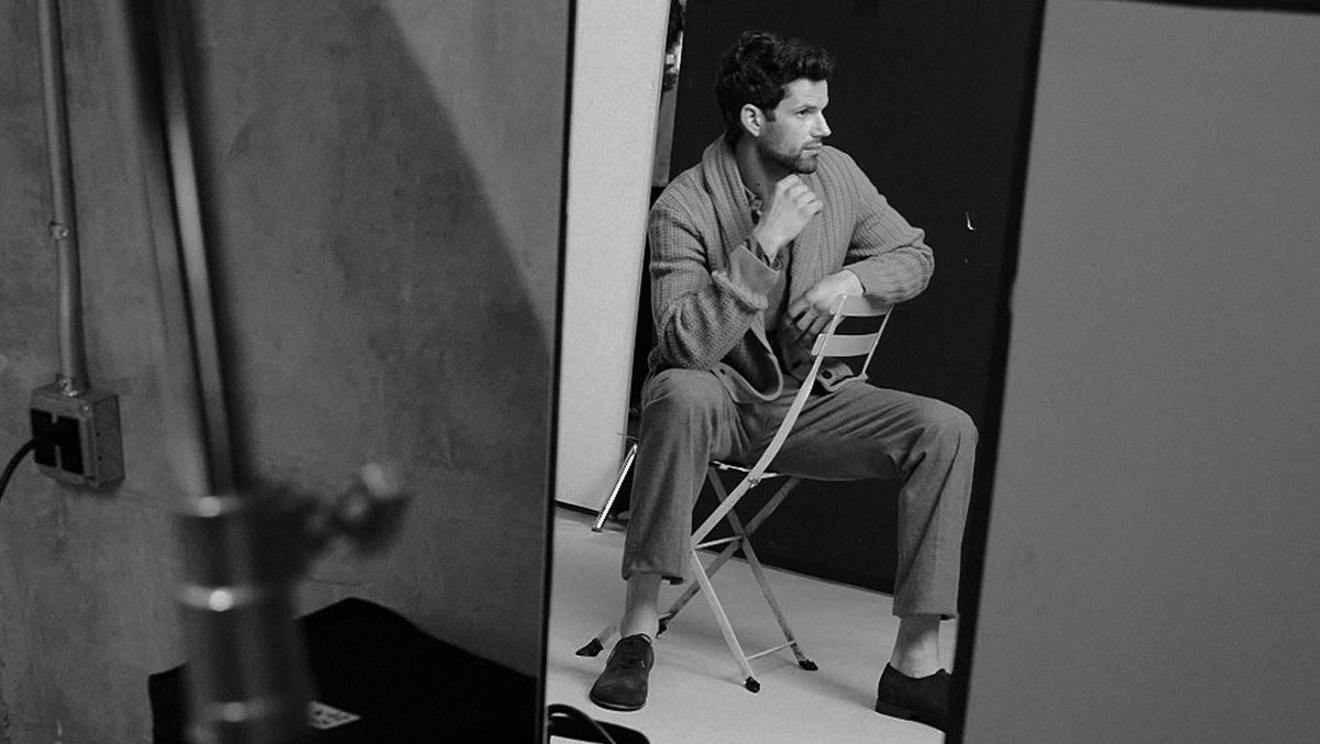 A black and white photograph of a man sitting on a folding chair in a studio setting, wearing a matching two-piece suit. The image is framed by a large mirror on the left, with studio lights and equipment visible in the foreground and background.