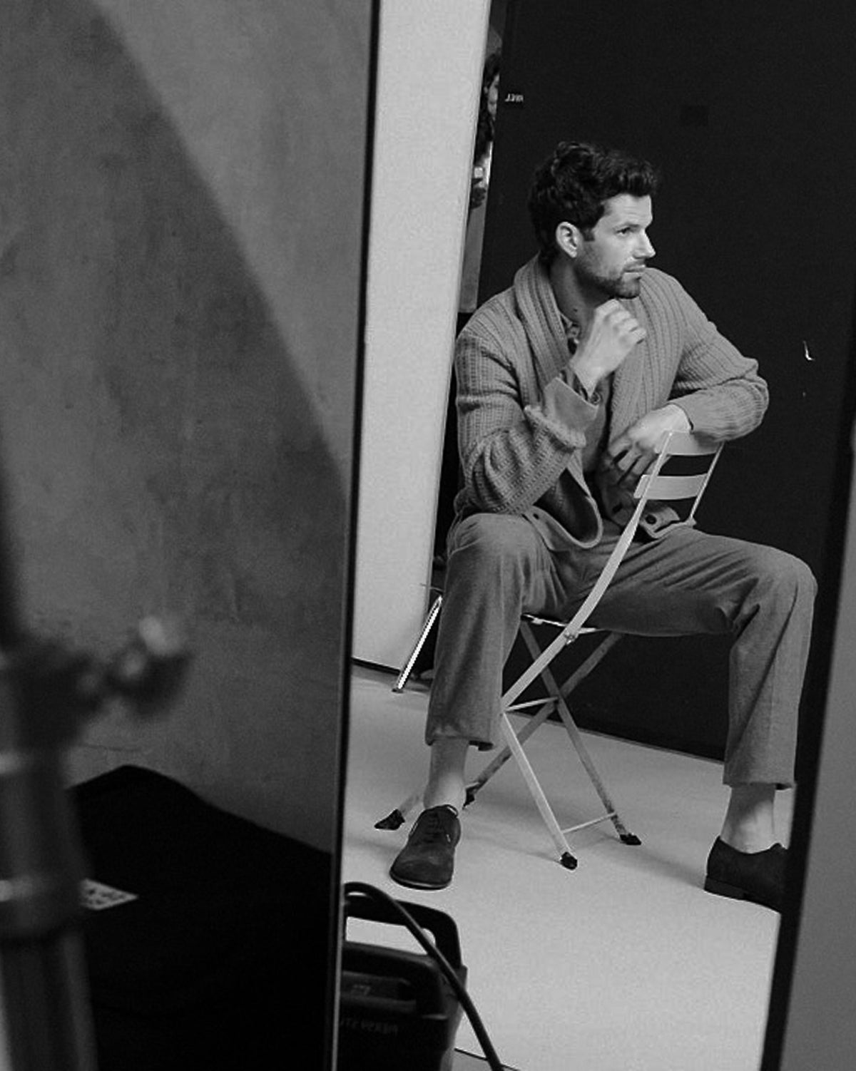 A black and white photograph showing a man sitting on a folding chair in a studio setting, wearing a light-colored cardigan and matching trousers.