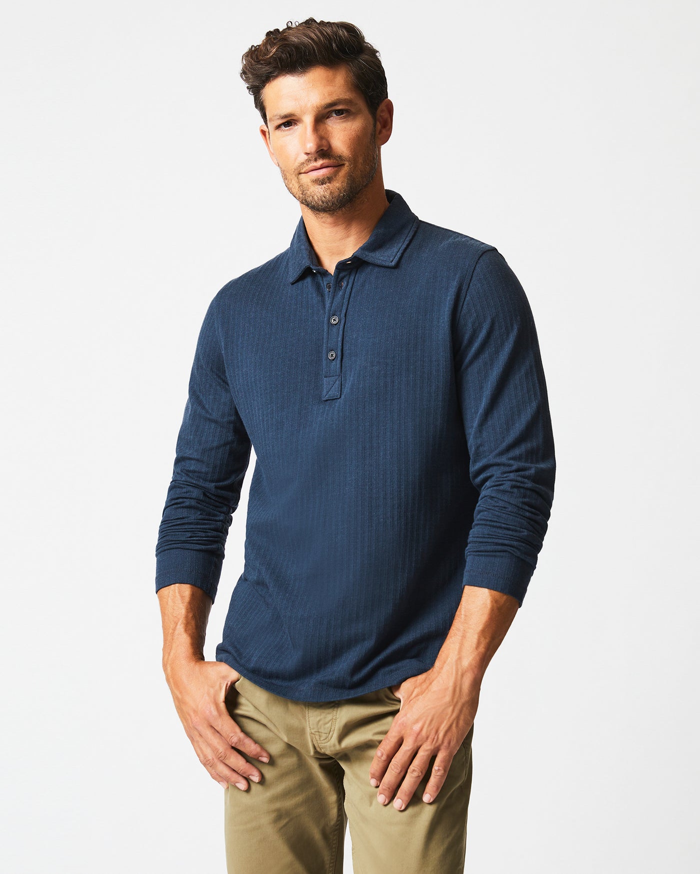 A man with short brown hair and a beard wears a navy blue long-sleeve polo shirt and khaki pants, standing against a plain white background with a slight smile and relaxed posture.