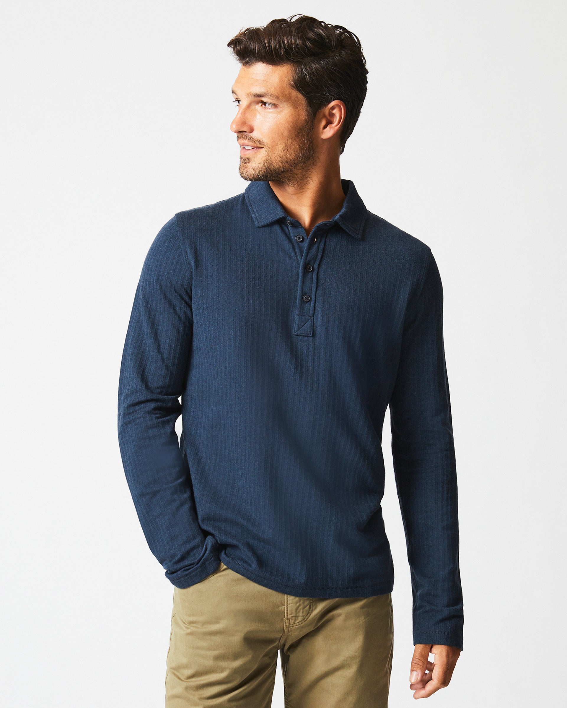 A man wearing a long-sleeve, dark blue polo shirt and khaki pants stands with one hand in his pocket, looking to his left against a plain white background.