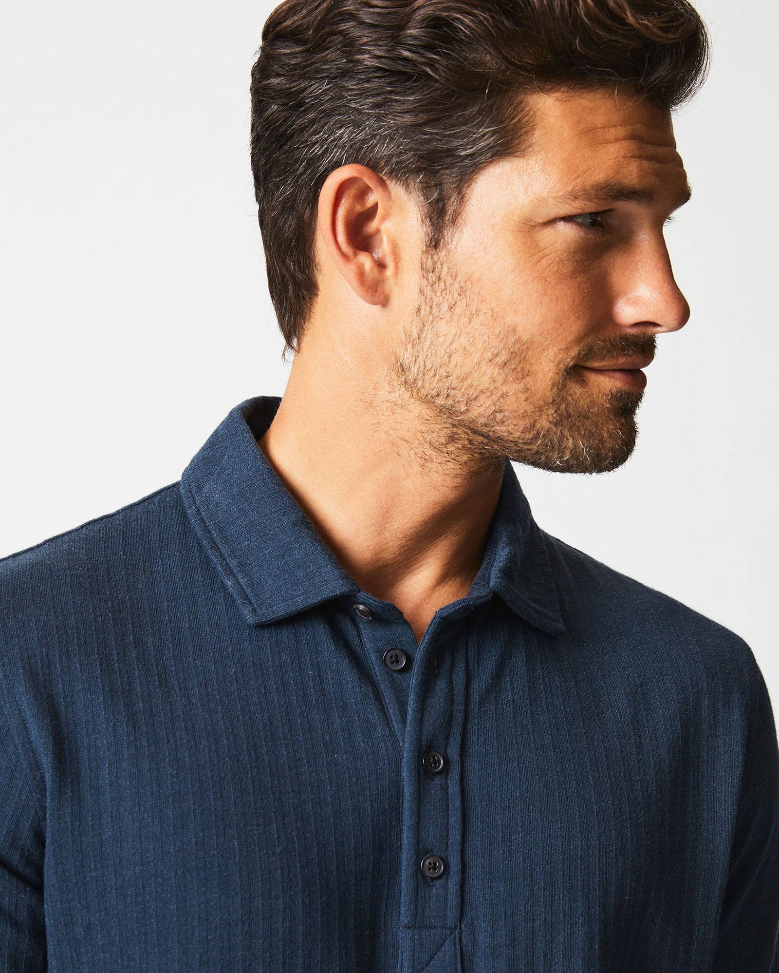 A man with short brown hair and a trimmed beard is wearing a dark blue collared, button-up shirt. He is looking to the right, and the background is plain white.