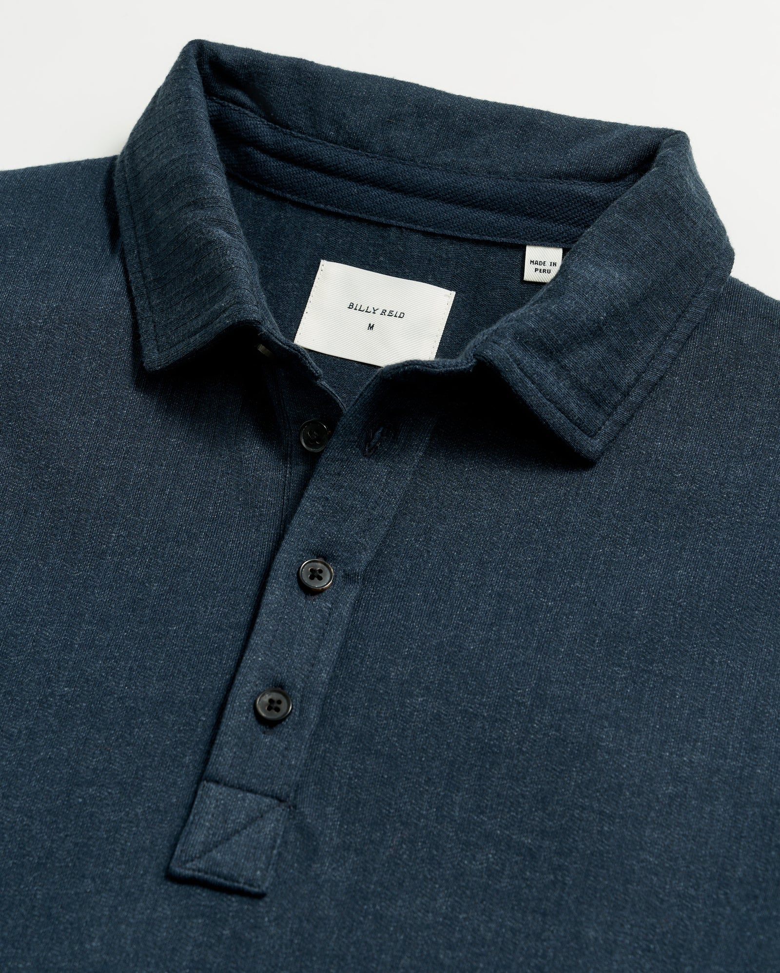 A close-up of a dark blue collared shirt with three black buttons and a visible white label inside the collar that reads BILLY REID M.