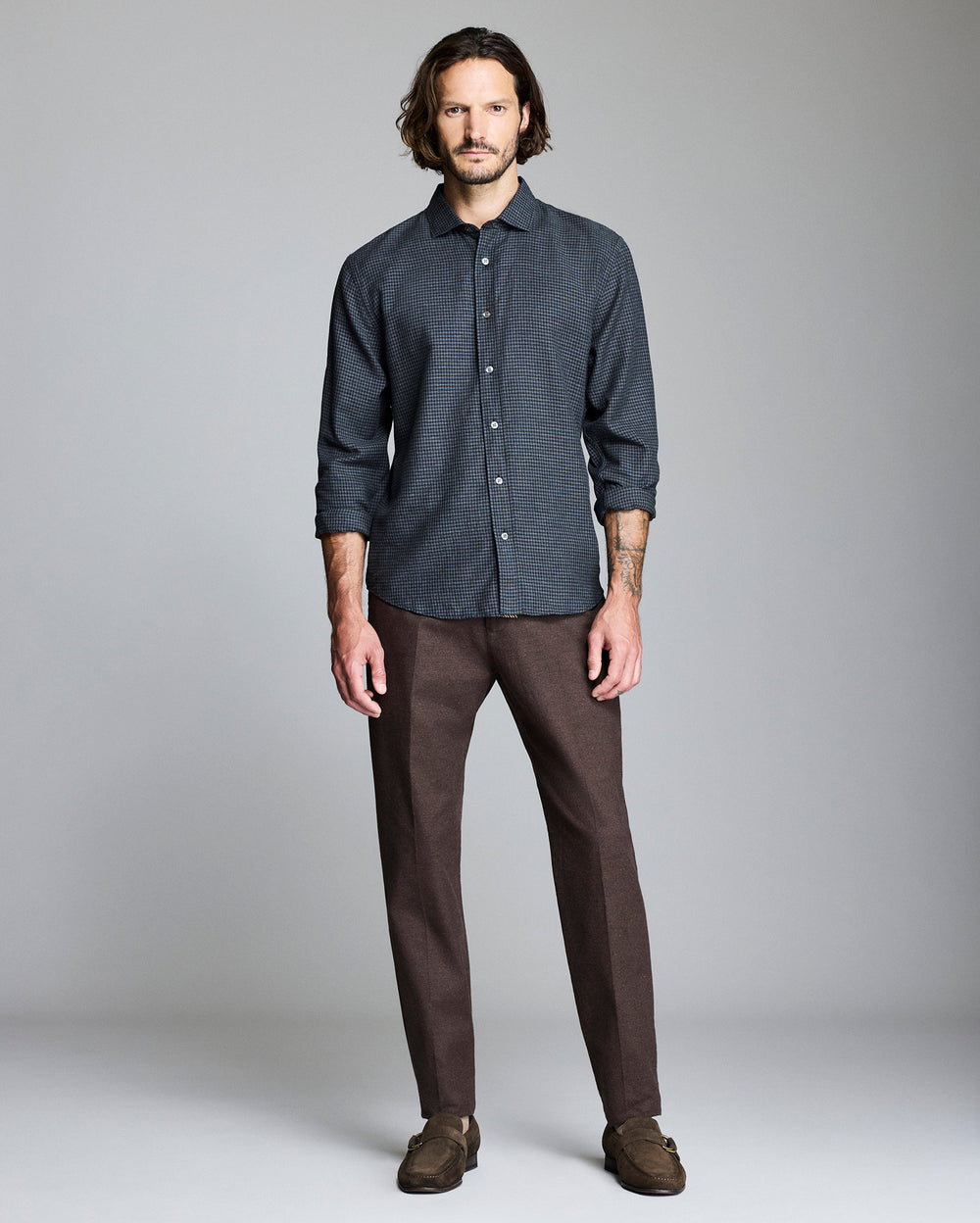 Man wearing a dark patterned shirt and brown pants against a gray background