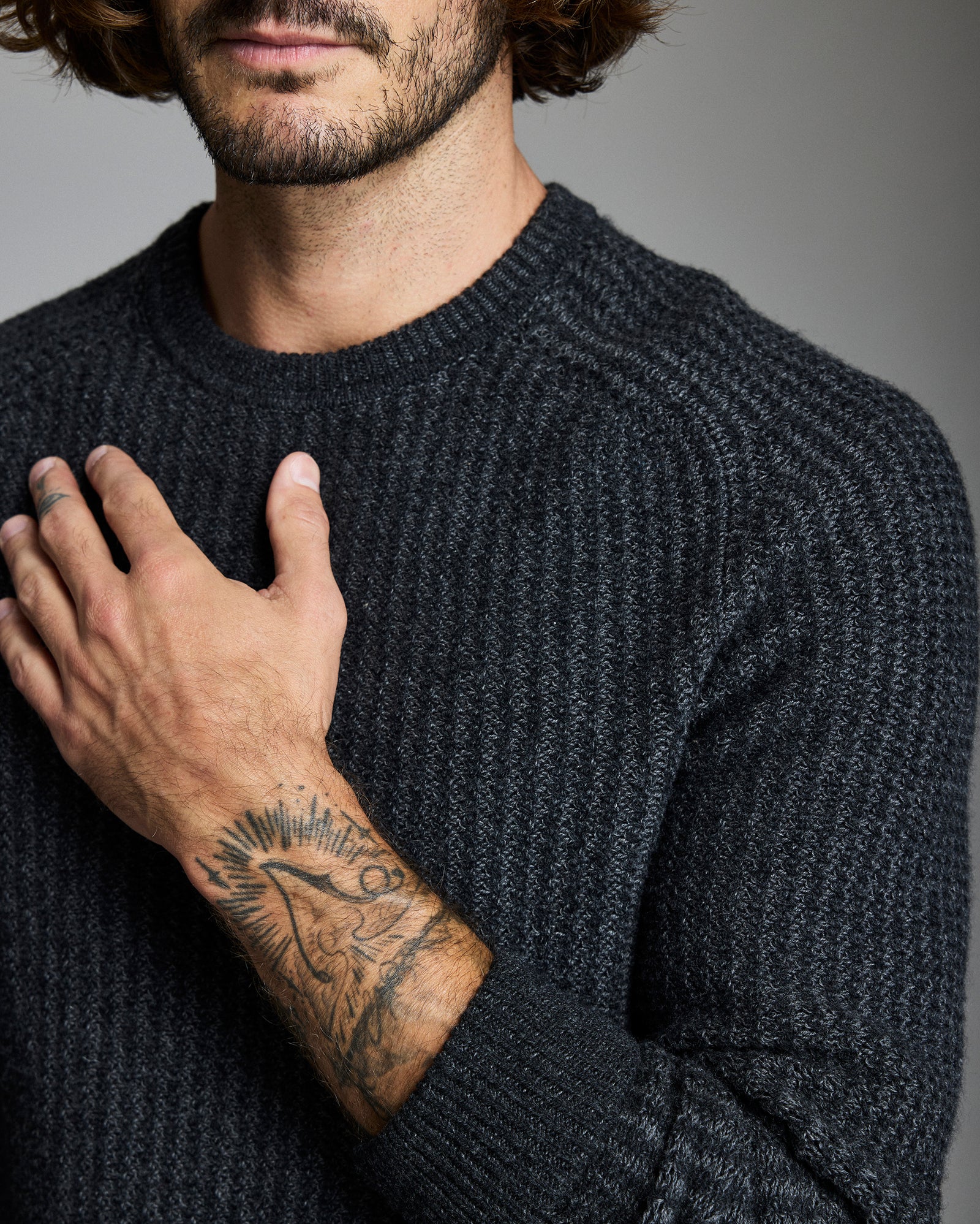 Man wearing a dark gray sweater with a tattoo on his arm, against a neutral background