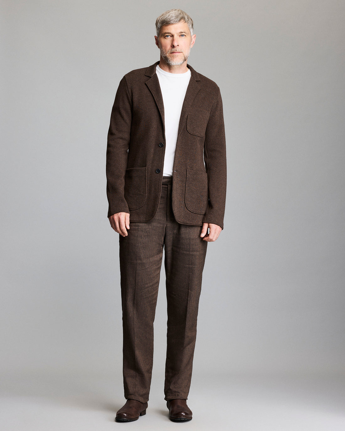 A man with short gray hair and beard wears a brown suit with a white t-shirt and brown shoes, standing against a plain light gray background.
