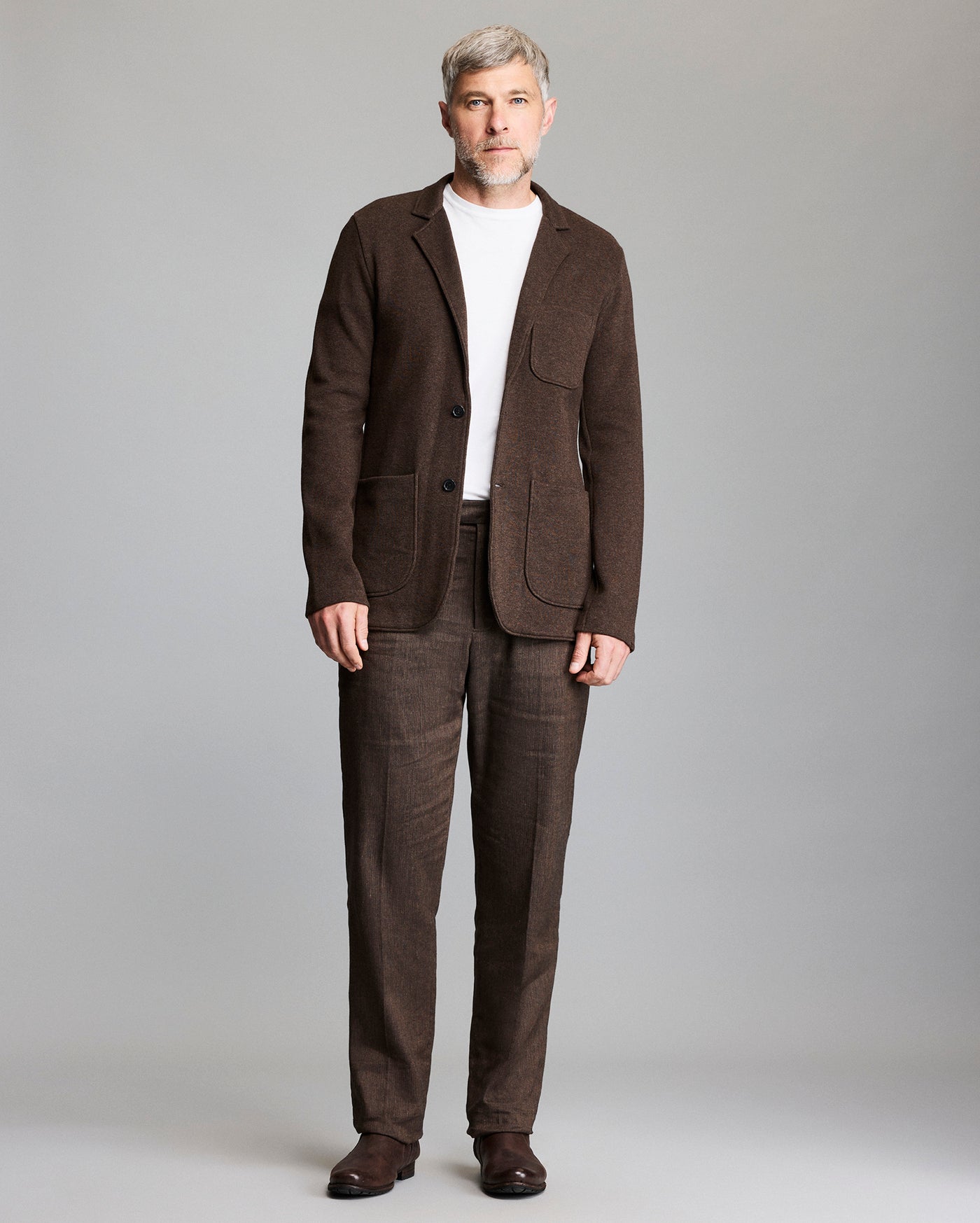 A man with short gray hair and beard wears a brown suit with a white t-shirt and brown shoes, standing against a plain light gray background.