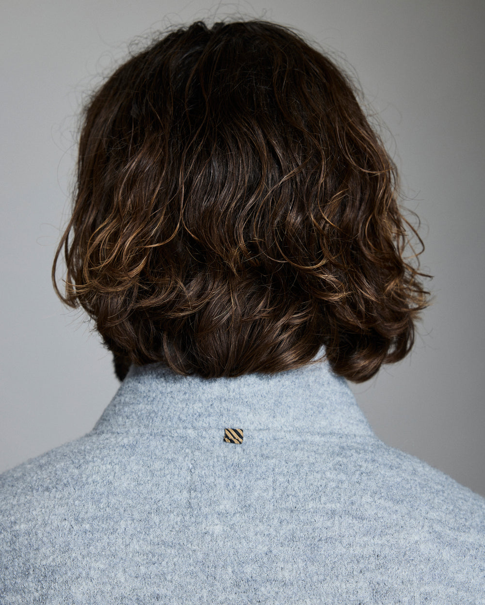 Back view of a person wearing a light gray coat with a brand logo on a plain background