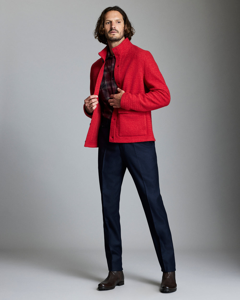 Man wearing a red jacket over a plaid shirt with navy pants against a gray background