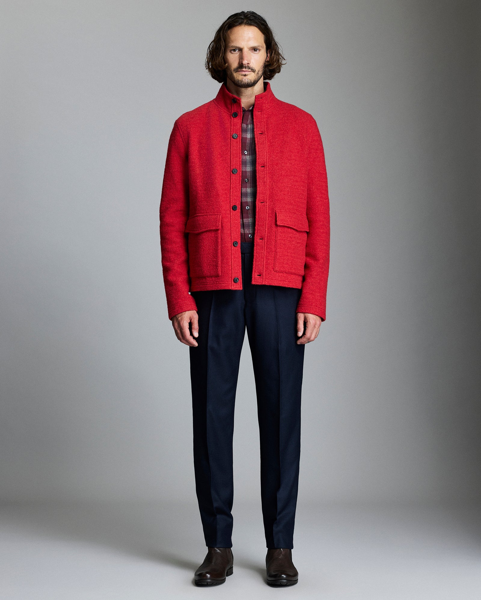 Man wearing a red jacket over a plaid shirt with navy pants against a gray background