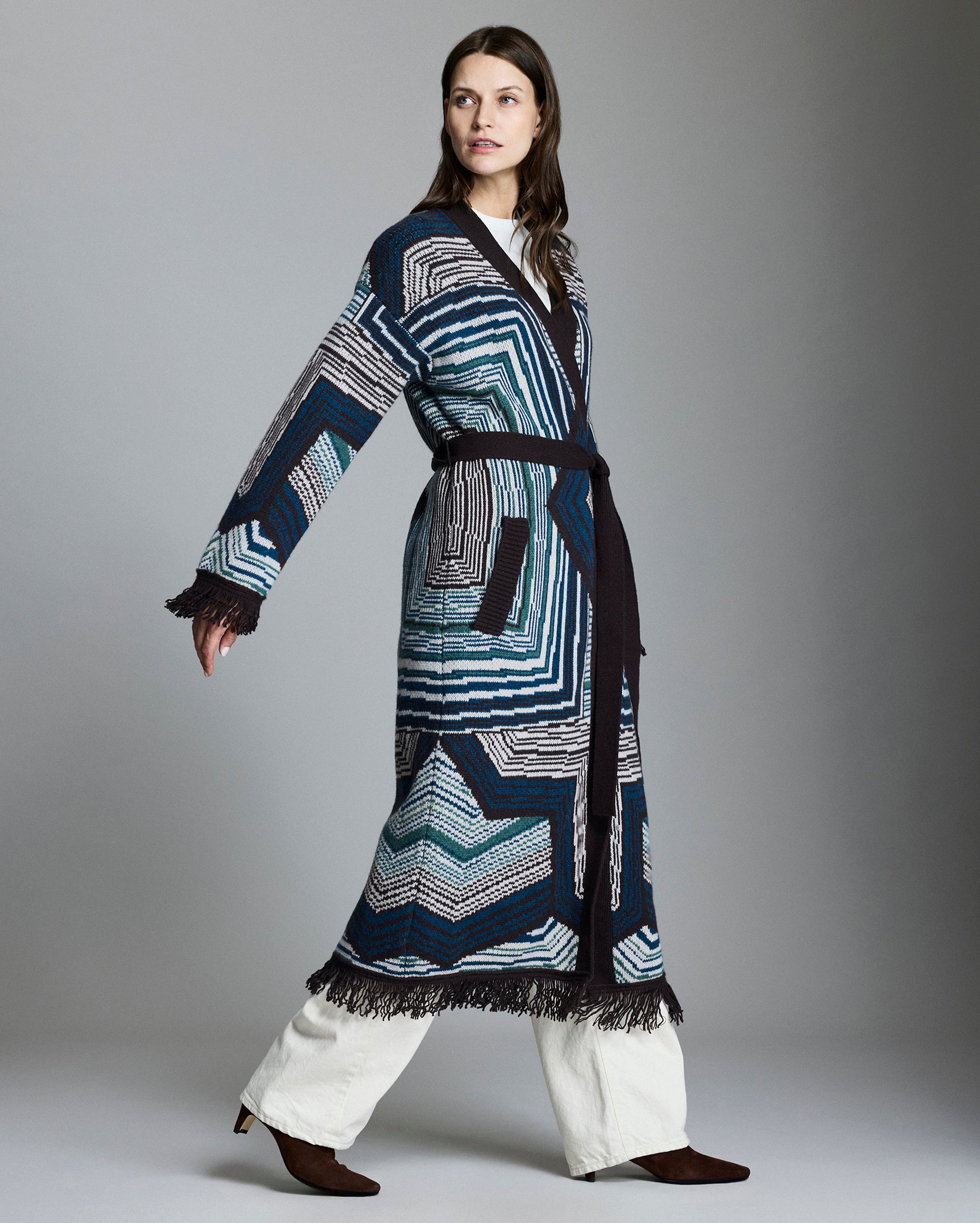 Woman wearing a long, patterned robe over white pants on a gray background