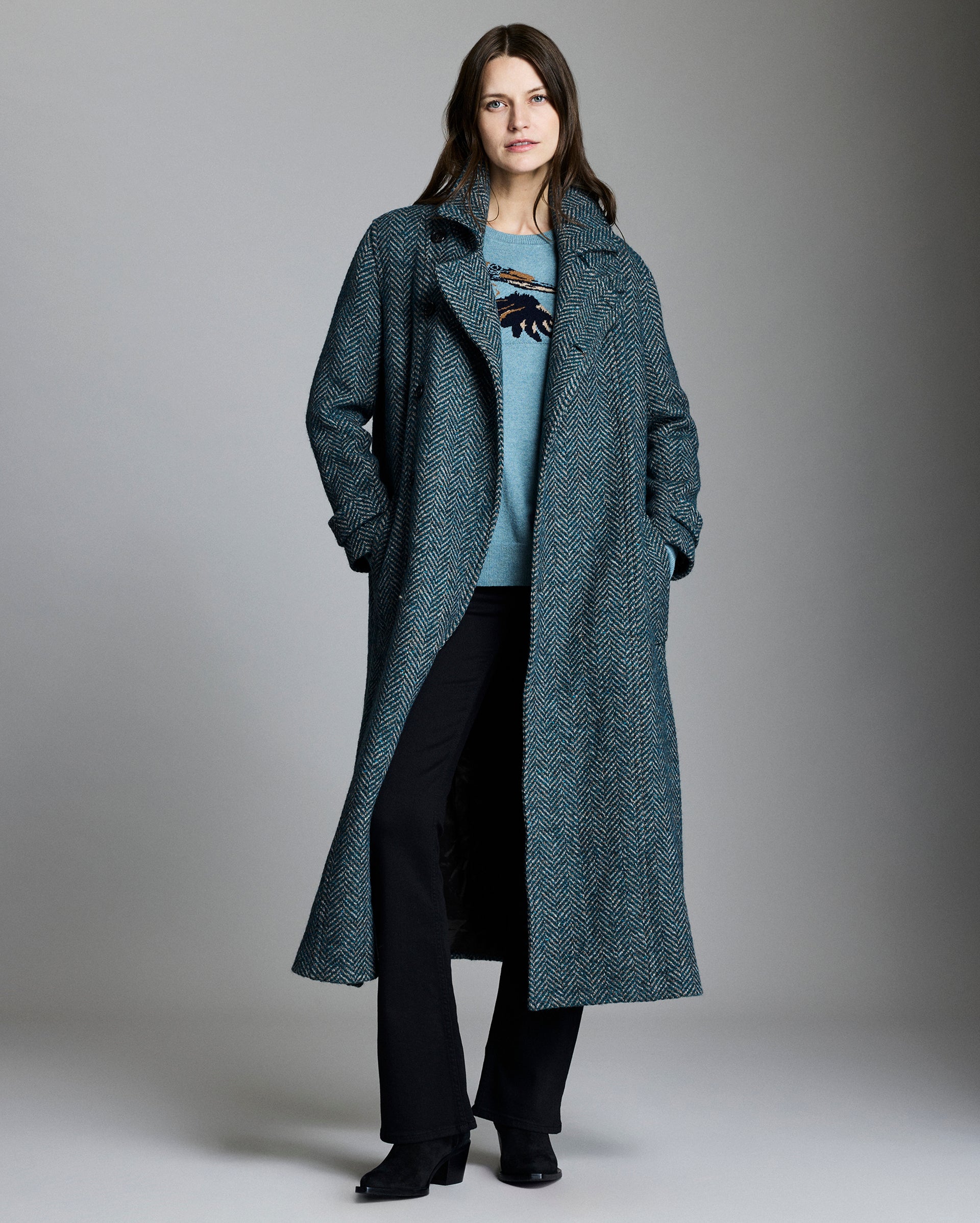Woman wearing a long green coat over a light blue sweater on a plain background
