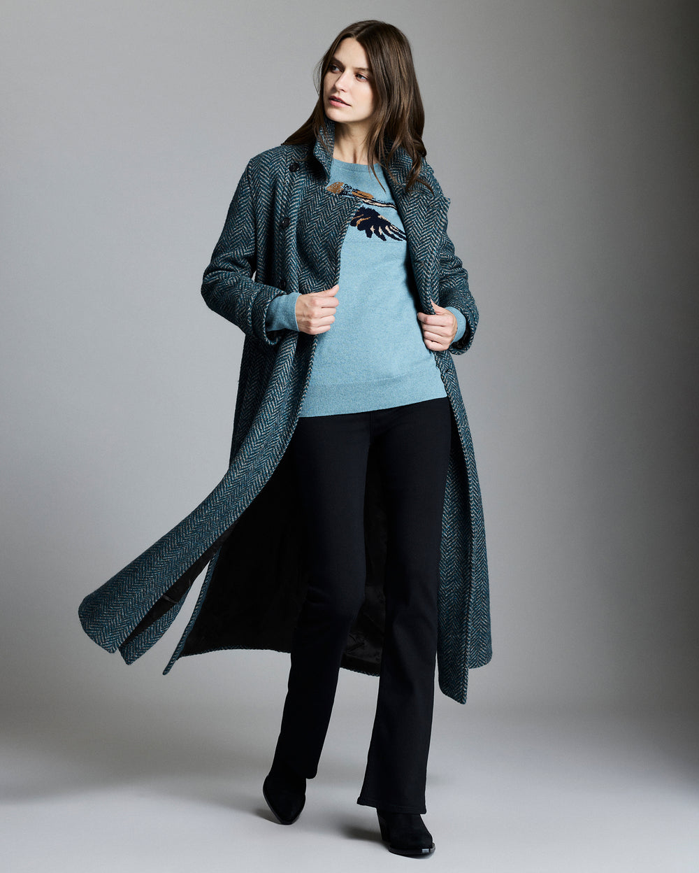 Woman wearing a long dark coat over a light sweater and black pants on a gray background