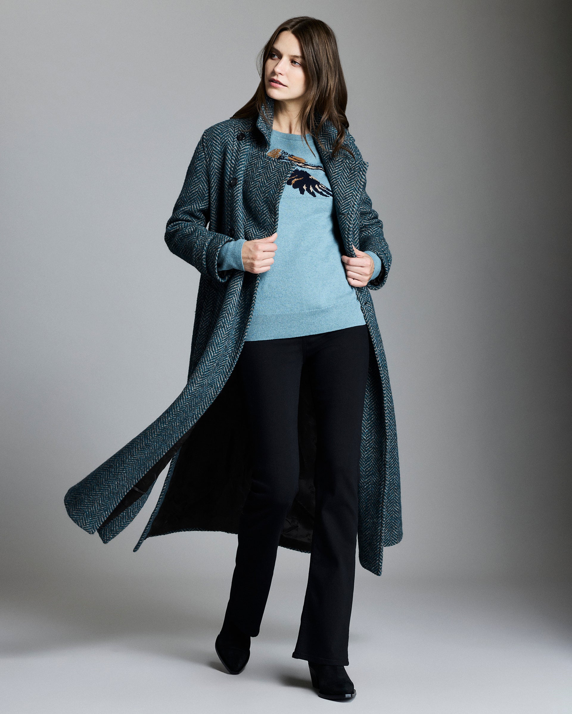 Woman wearing a long dark coat over a light sweater and black pants on a gray background