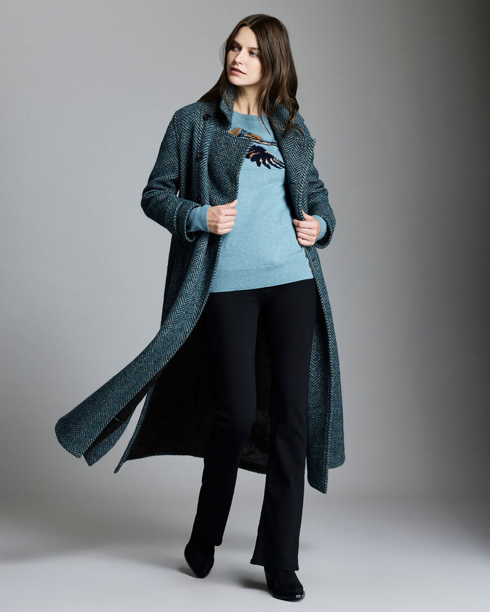 Woman wearing a long dark coat over a light sweater and black pants on a gray background