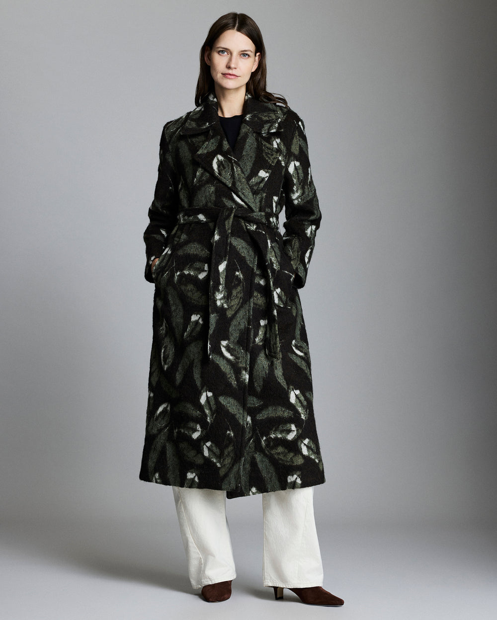 Woman wearing a black patterned coat with white pants on a gray background