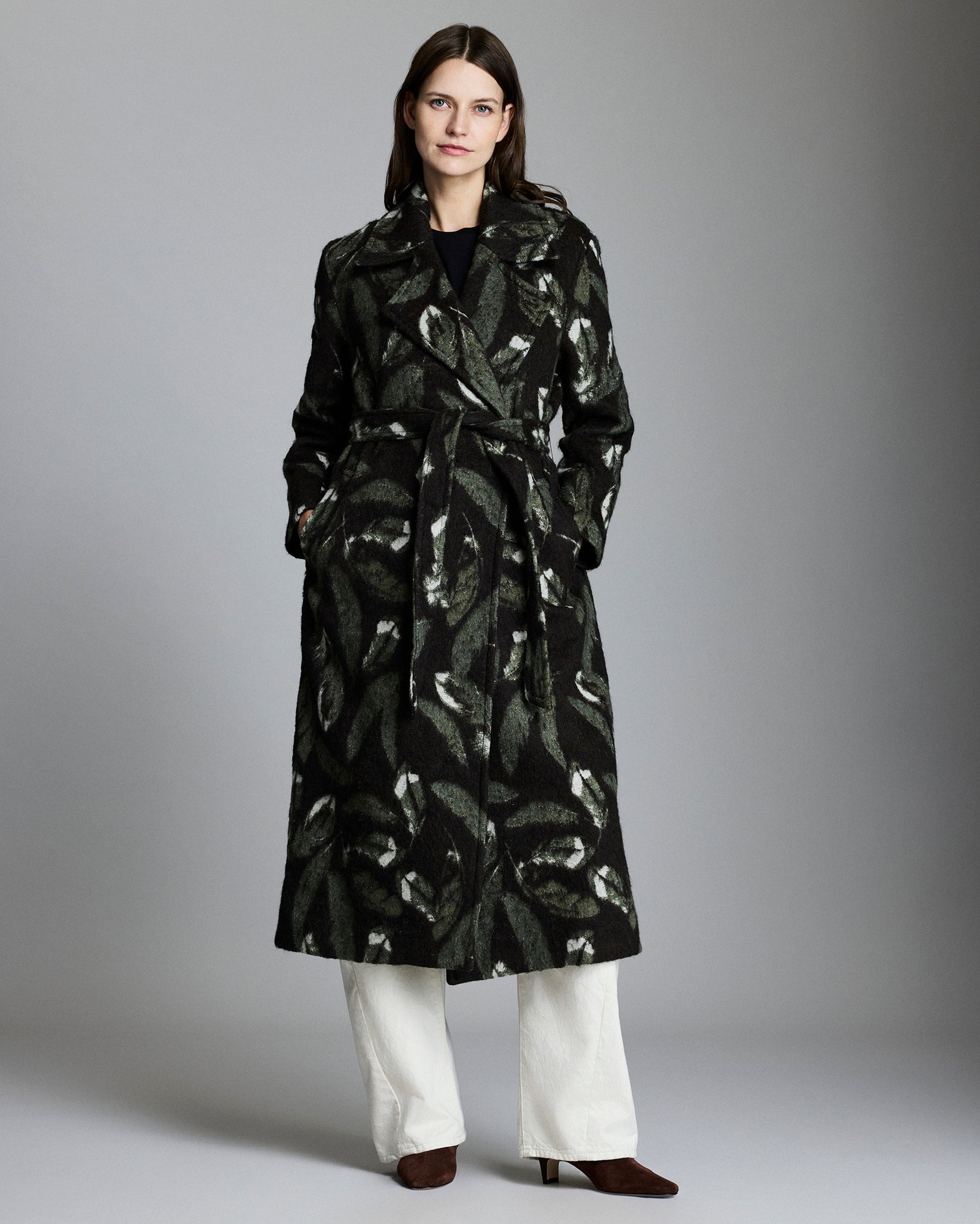 Woman wearing a black patterned coat with white pants on a gray background