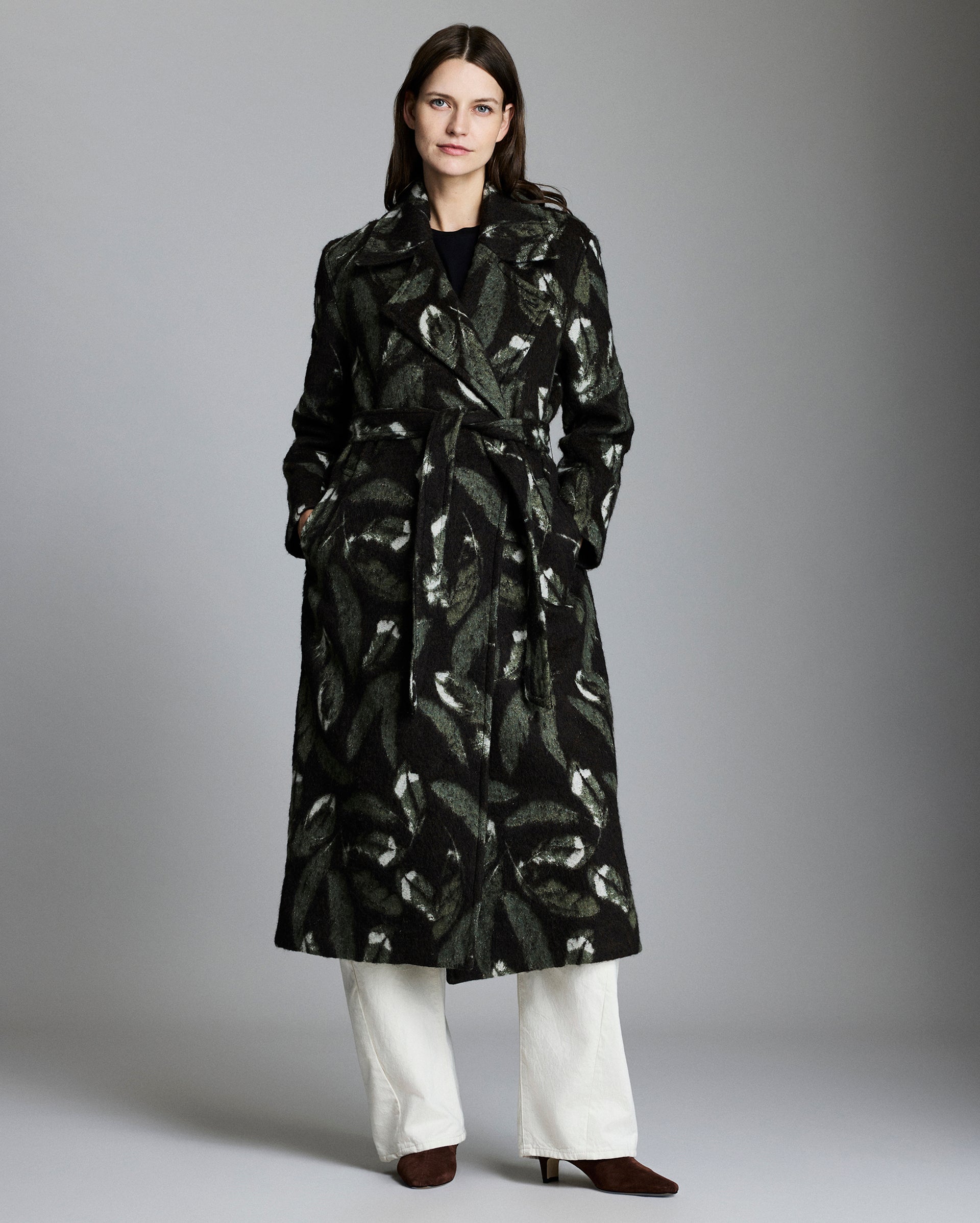Woman wearing a black patterned coat with white pants on a gray background