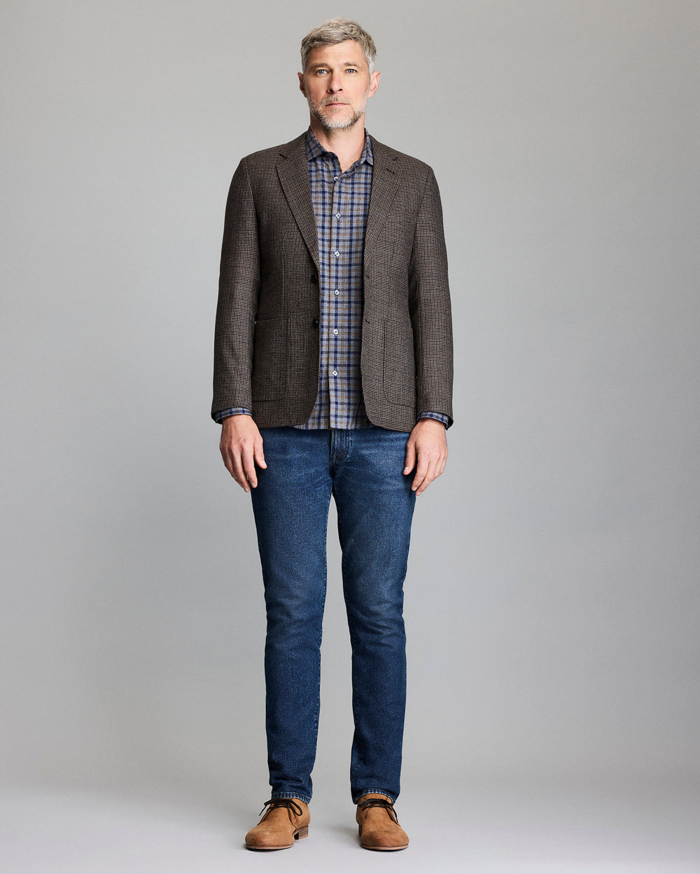 Man wearing a brown checkered blazer, plaid shirt, blue jeans, and brown shoes on a gray background