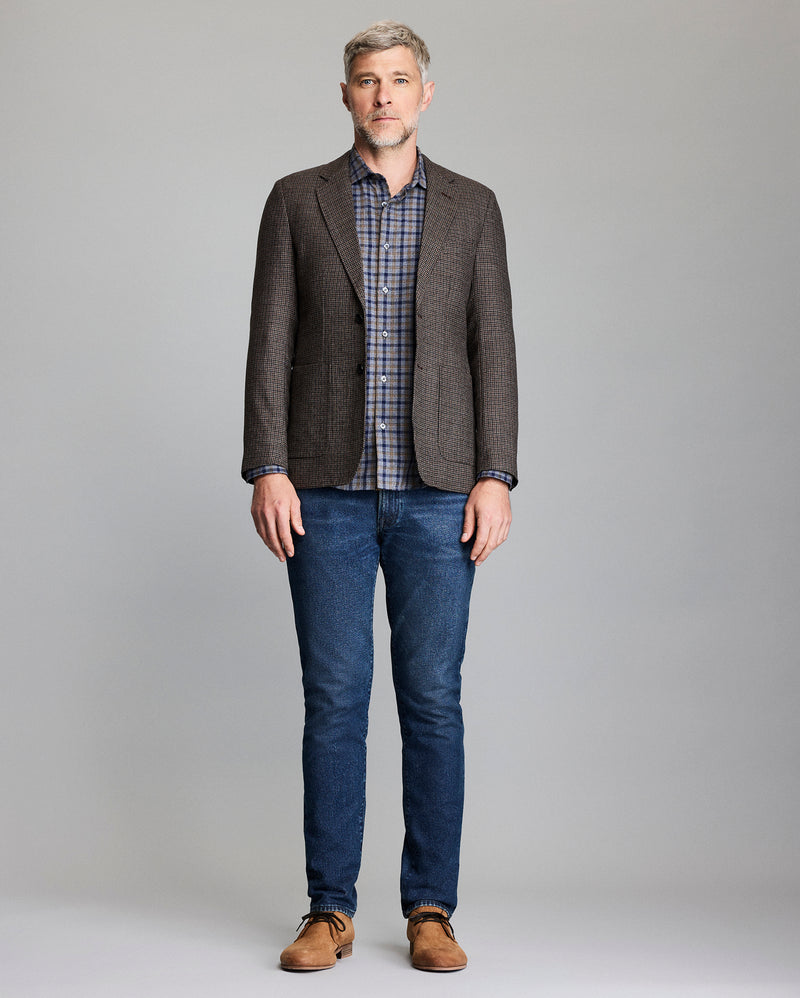 Man wearing a brown checkered blazer, plaid shirt, blue jeans, and brown shoes on a gray background Alternate