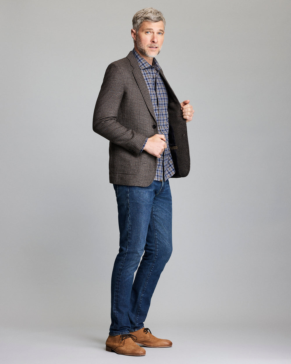 Man wearing a gray blazer, plaid shirt, blue jeans, and brown shoes on a gray background