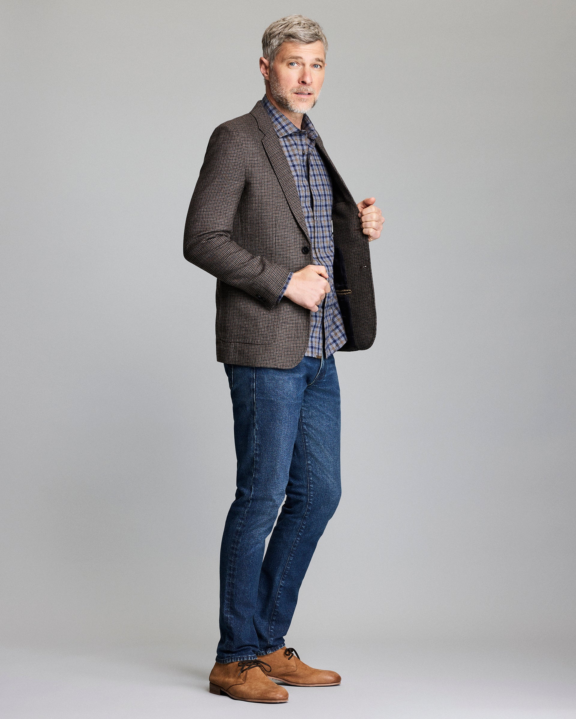 Man wearing a gray blazer, plaid shirt, blue jeans, and brown shoes on a gray background