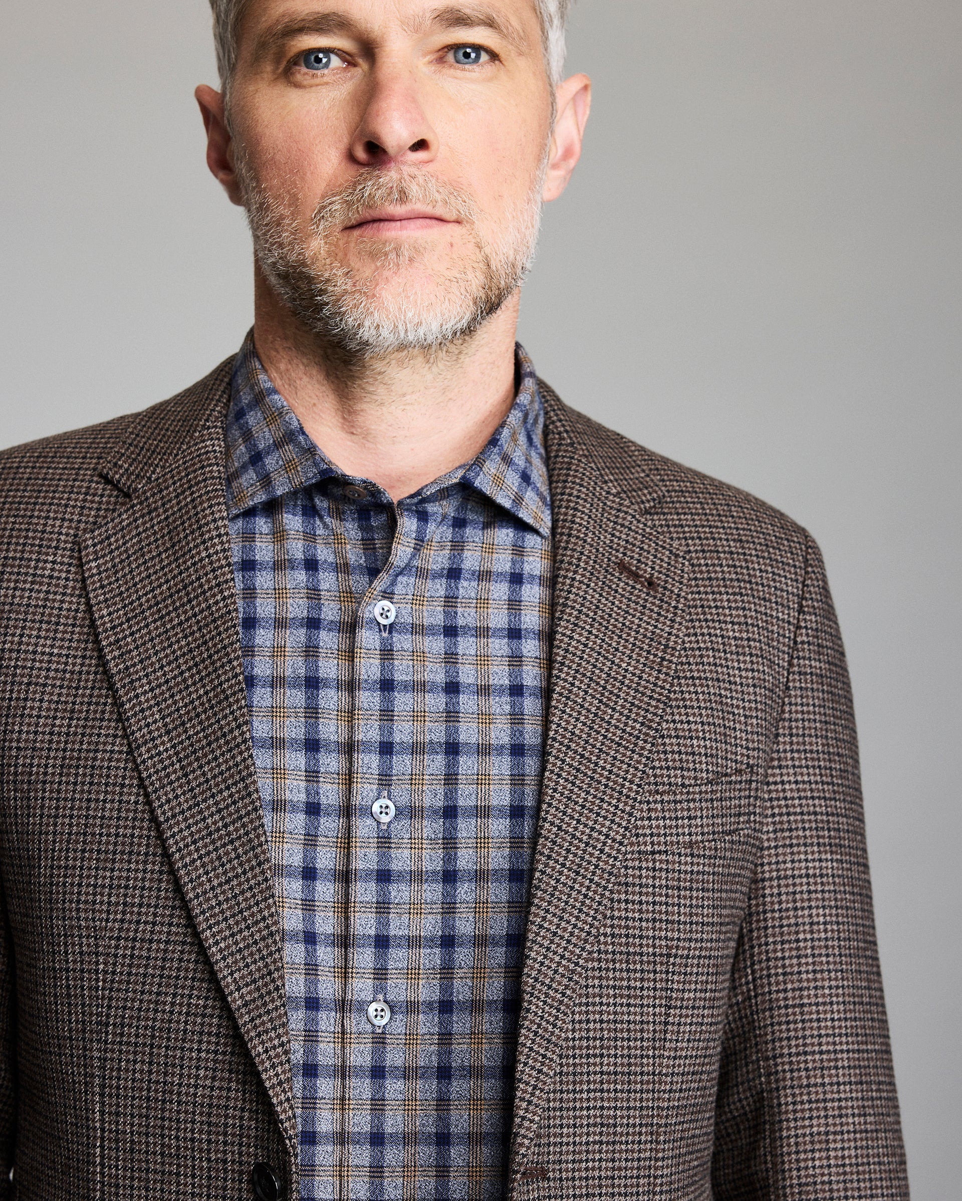 Man wearing a brown checkered blazer over a plaid shirt against a gray background