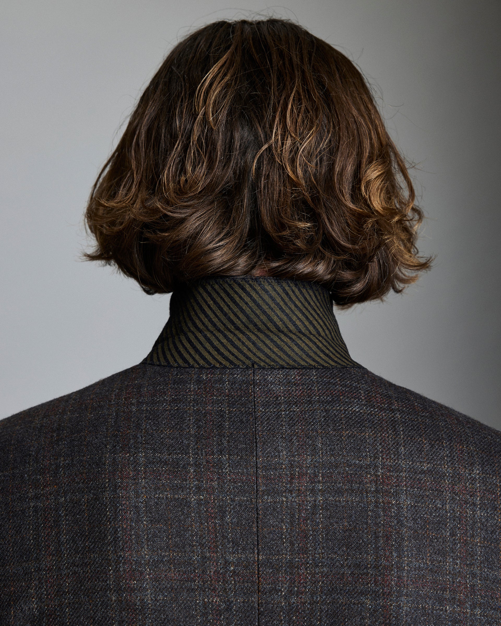 Person wearing a dark gray checkered suit with a textured shirt and wavy brown hair.