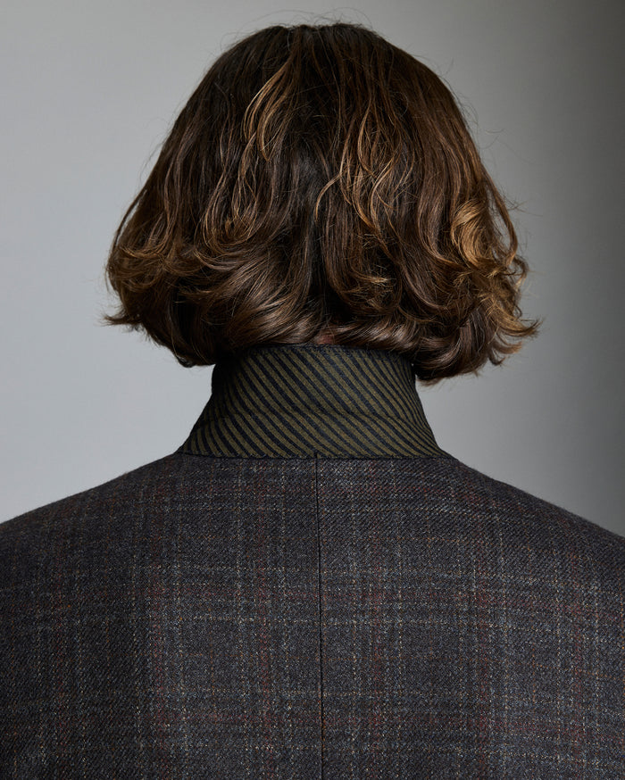 Person wearing a dark gray checkered suit with a textured shirt and wavy brown hair.